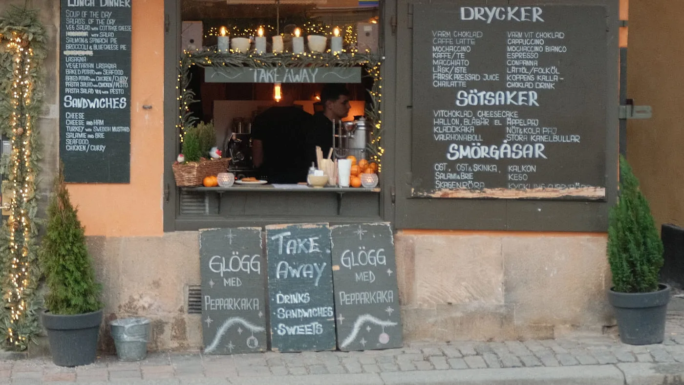 Kafé mot gatan med öppen serveringslucka, krittavlor med meny och granar i kruka utanför.
