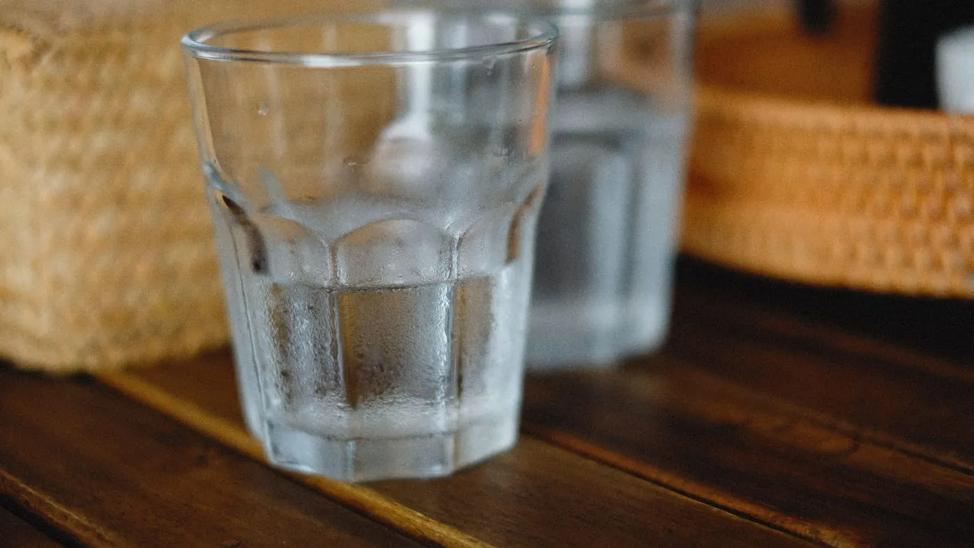 Ett glas kallt vatten med kondens på utsidan, på ett mörkt träbord bredvid en brödkorg.