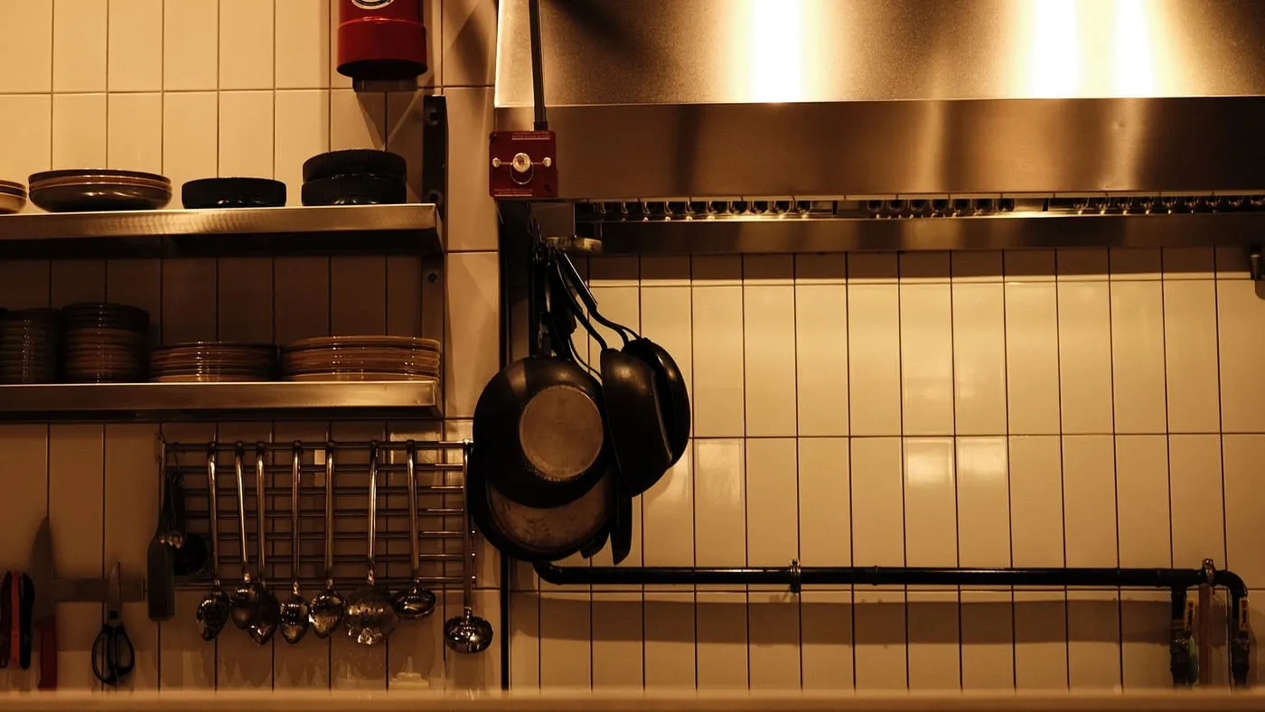 Kaklad vägg i ett tomt restaurangkök med hyllor för tallrikar, upphängda stekpannor och slevar under en stålkåpa.