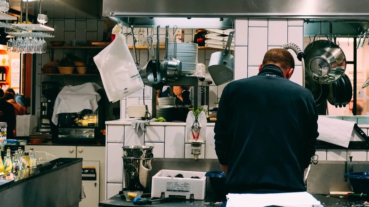 Kock med ryggen mot kameran arbetar vid passet i ett kommersiellt kök med vitt kakel, hängande kastruller och stålkåpa.