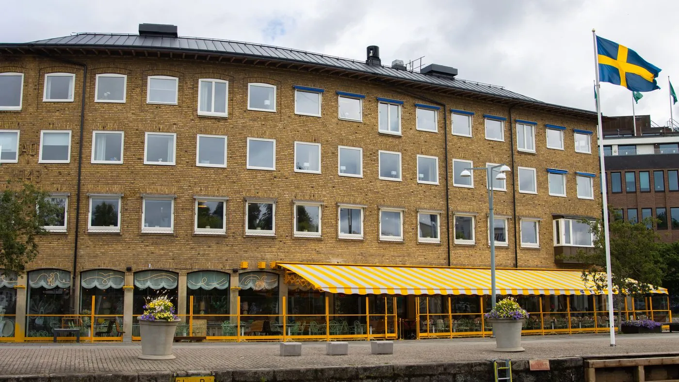 Gult tegelhus vid en kaj med en restaurang bakom gulvit markis och en svensk flagga intill.
